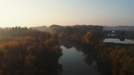 Aerial-shot-of-morning-sunrise-with-light-rays-breaking-through-fog-over-Danube-river-and-Colorful-Autumn-forest