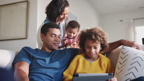 Dad-and-daughter-on-sofa-using-tablet,-mum-behind-them-holding-toddler,-low-angle,-close-up