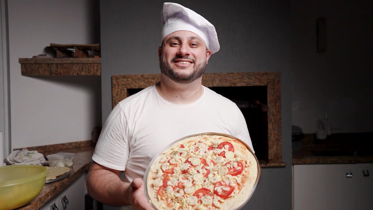 Uno chef professionista sorride alla telecamera mentre tiene orgogliosamente una pizza appena preparata, pronta per essere infornata