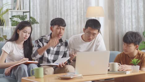 asian teen group studying on a laptop at home. taking online course, a boy in plaid shirt hands up, asking question, giving an answer, writing into notebook