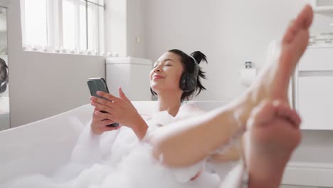 Video-of-portrait-of-smiling-biracial-woman-with-headphones-and-smartphone-in-bathtub-in-bubble-bath