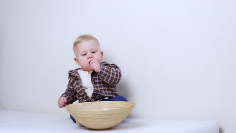 adorable blond baby in checkered jacket sits at the wooden bowl. kid holds a pebble at his mouth. white backdrop.