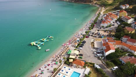 Playa-De-Baska,-Isla-De-Krk,-Croacia---Vista-Aérea-De-Drones-Del-Balneario-Con-Costa,-Turistas,-Bulevar,-Tumbonas-Y-Parque-Acuático