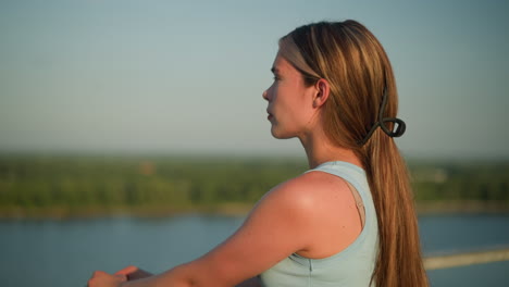 white lady standing outdoors with hands brought forward, hair tied back, gazing into distance with a thoughtful expression, under warm sunlight, background is blurred with natural scenery