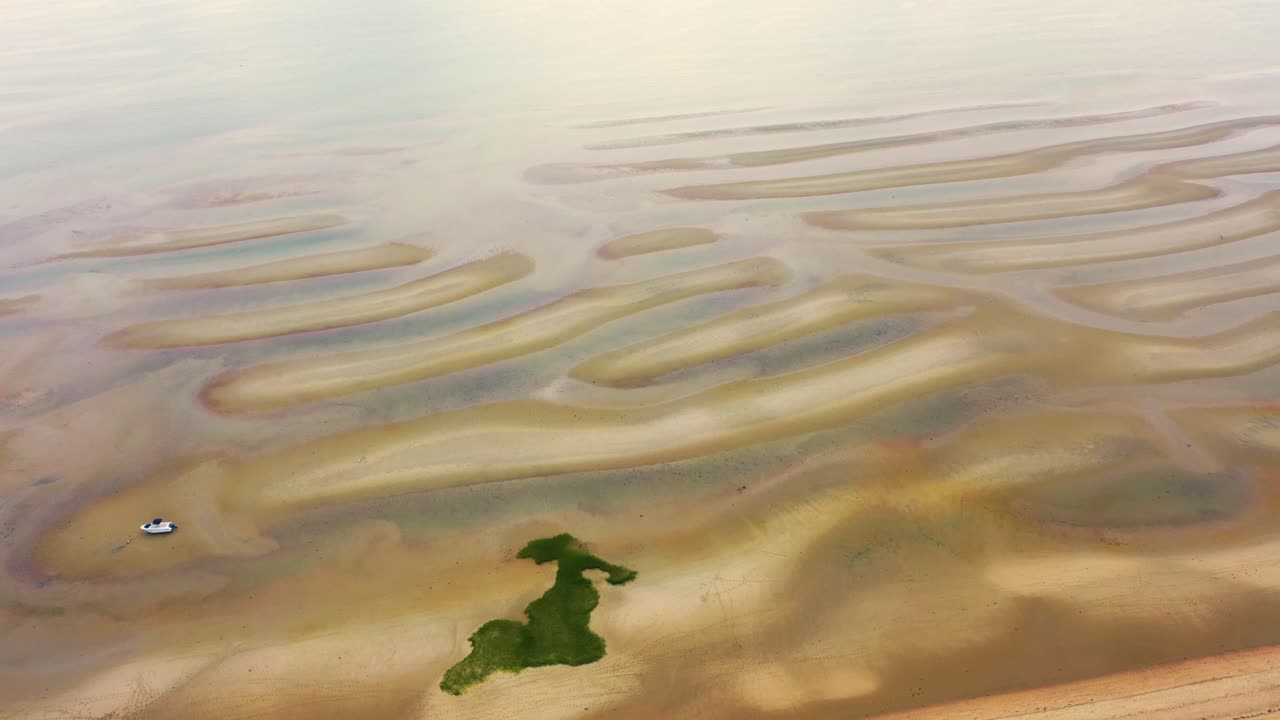 Drone captures a wide sandy beach at low tide with dune-like ridges stretching outward, tide pools shimmering in shallow depressions, and expansive shoreline glowing under soft skies