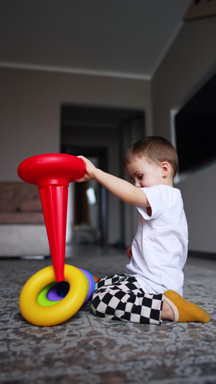 Two year old toddler sitting on the floor disassembling the pyramid. Baby boy plays with toys at home. Low angle view. Vertical video.