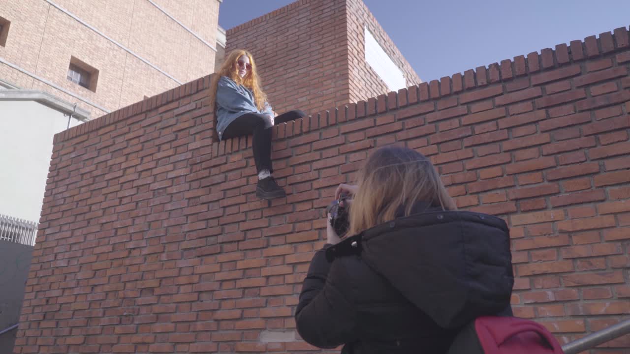 Two friends have fun taking pictures in the street. Redhead model poses sitting on top of a brick wall and smiles for her photographer friend