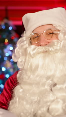 santa claus smiling on camera indoors. portrait of a real santa with white beard, in glasses and red costume standing in the living room on christmas tree background. vertical video