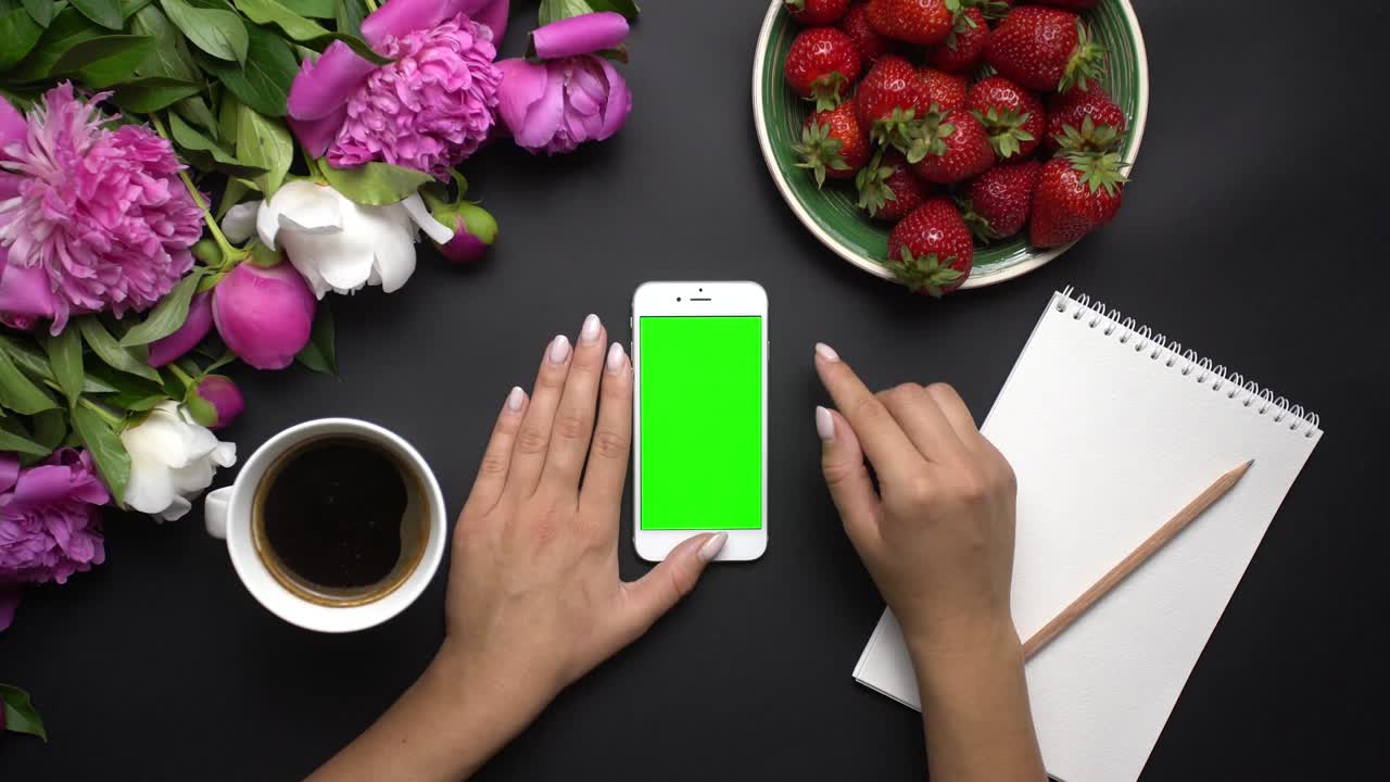 mujer usando dispositivo de teléfono inteligente blanco con pantalla verde. hermosas peonías blancas y rosas flores, fresas rojas, café y cuaderno de notas en el fondo negro. vista de la mesa. clave de croma