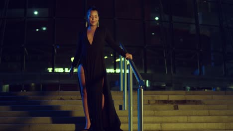 dama sexy con un vestido negro caminando por una escalera de un edificio de cristal por la noche en la ciudad