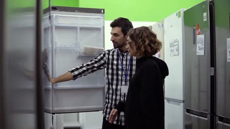Young-woman-standing-in-front-the-open-door-of-refrigerator-with-male-consultant-discussing-design-and-quality-before-buying-in