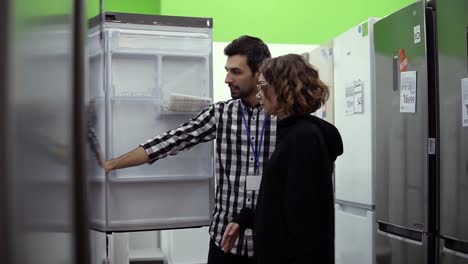 young woman standing in front the open door of refrigerator with male consultant discussing design and quality before buying in