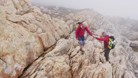 senior hiker couple with backpacks and trekking poles holding each others hands while climbing rocks