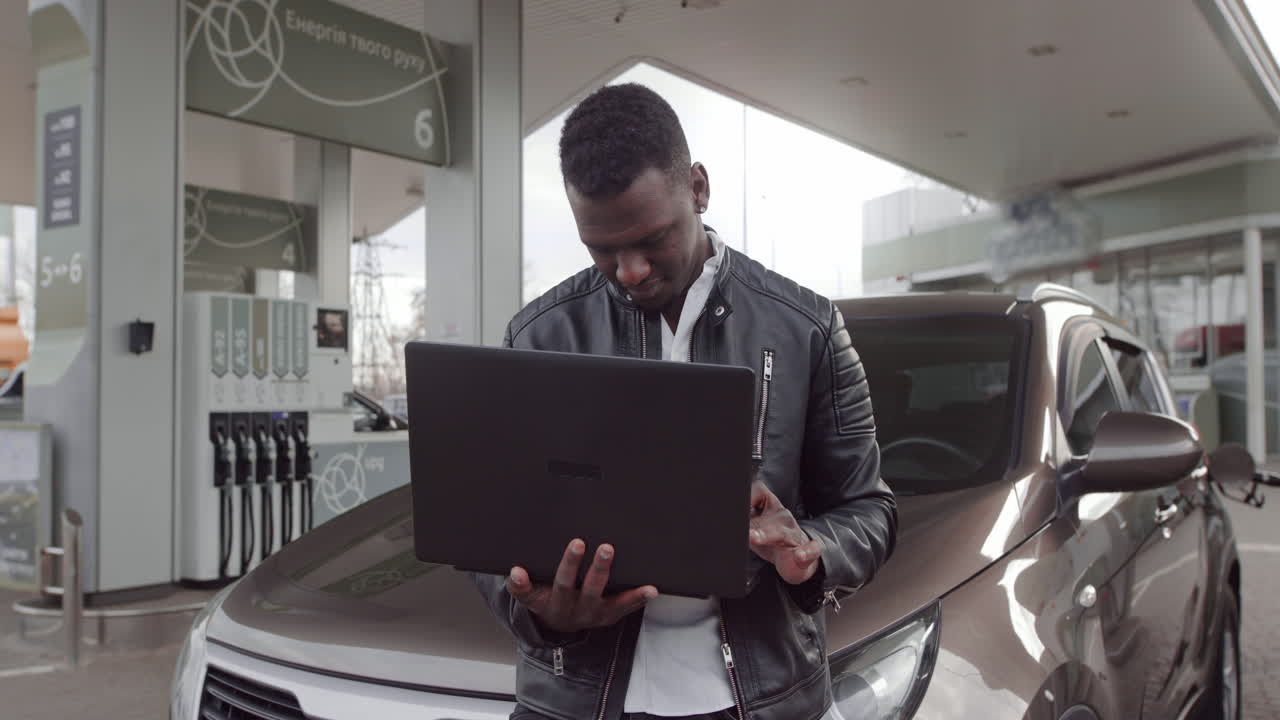 Black businessman at petrol station standing near the car with take away coffee and laptop pc and