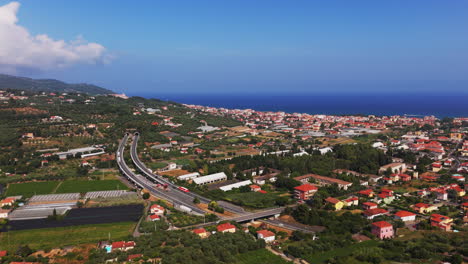 side drone shot in varigotti showing a road turning into a tunnel, with a bridge, farmland, vegetation, a coastal town and the sea