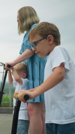 a mother is riding a scooter with her younger son, while the older child rides his scooter independently beside them