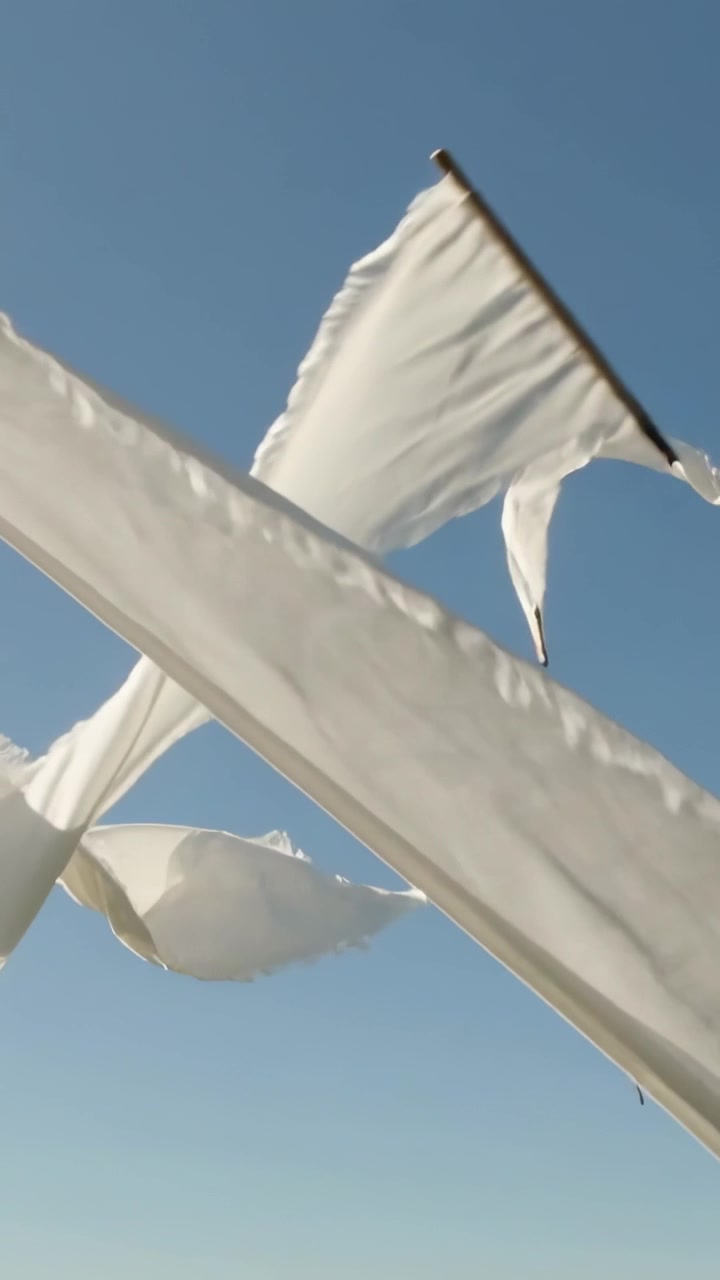 Video verticale: Biancheria bianca che ondeggia e volteggia sui tetti sotto il cielo blu a causa del vento rafficato, fissata con mollette.