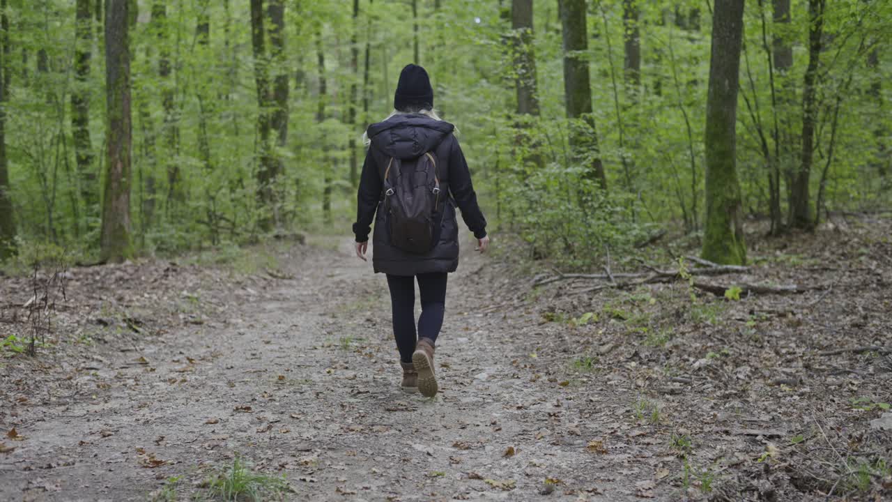 A woman wearing a dark jacket and backpack walks alone along a forest path surrounded by tall green trees. The peaceful scene captures the calm and solitude of a nature hike