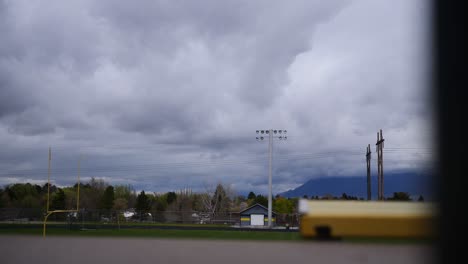 Storm-clouds-rolling-over-American-football-track-and-field-on-cloudy-rainy-day,-with-green-trees-and-mountains-in-background,-and-yellow-goal-post-and-stands