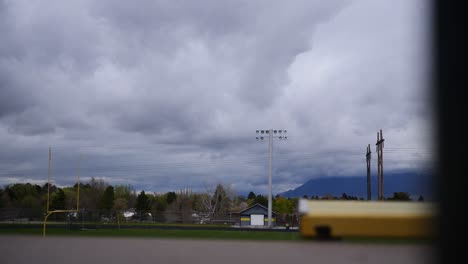 nubes rodando sobre la pista y el campo de fútbol americano en un día nublado y lluvioso, con árboles verdes y montañas en el fondo, y postes de gol amarillos y puestos