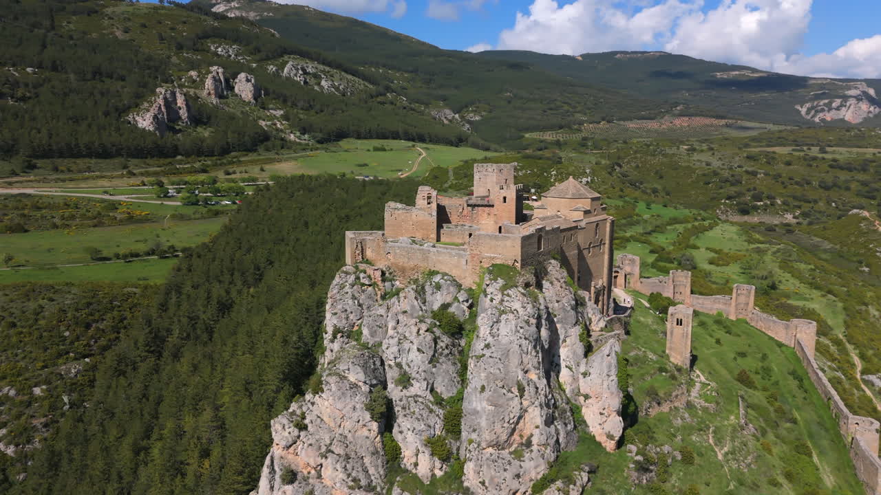 Drone orbiting an ancient stone castle perched on a rocky cliff, with panoramic views of green hills and forests under a blue sky. Captured in high-resolution during daylight