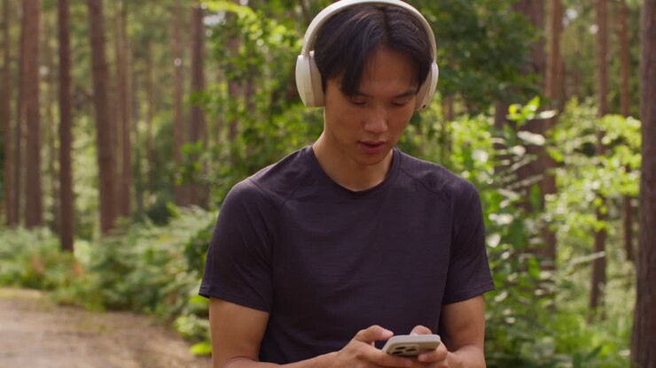 Uomo con cuffie wireless che si allena all'aperto, ascoltando musica o podcast in streaming dal cellulare, correndo lungo un sentiero nel bosco e indossando abbigliamento sportivo