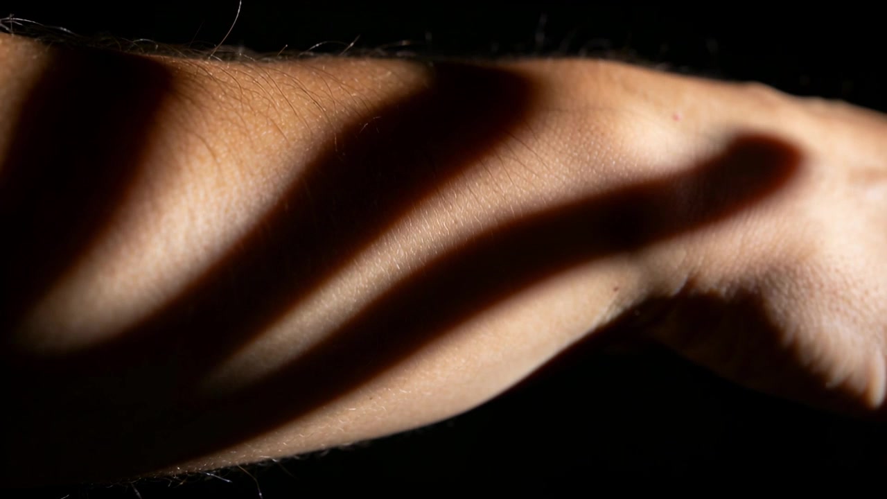 Una mano che si muove sopra un avambraccio nudo, fasce d'ombra che scivolano verso il polso in studio sotto la luce, con un cavo
