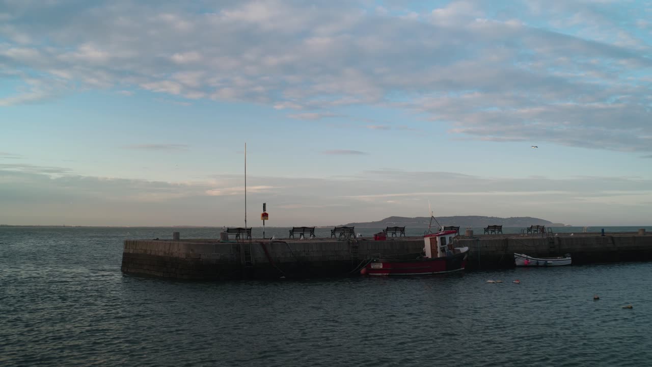 A locked-off shot of Bullock Harbour in Dublin, Ireland. A fishing boat is moored at the stone pier with calm waters, benches, and a coastal view under a soft blue sky.