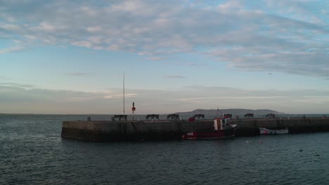 a locked-off shot of bullock harbour in dublin, ireland. a fishing boat is moored at the stone pier with calm waters, benches, and a coastal view under a soft blue sky.