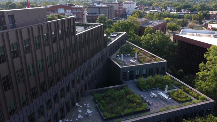 Fachada escalonada y patios con vegetación de un edificio de uso mixto de ladrillo oscuro en Charlottesville, Virginia, con los tejados del centro y colinas brumosas al fondo. Toma aérea de aproximación