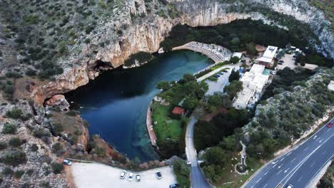 Panorámica-A-Vista-De-Pájaro-Del-Lago-Sumergido-Vouliagmeni-En-Atenas,-Grecia,-Con-Impresionantes-Acantilados-Circundantes-Y-La-Carretera-Sobre-El-Lago-|-4k