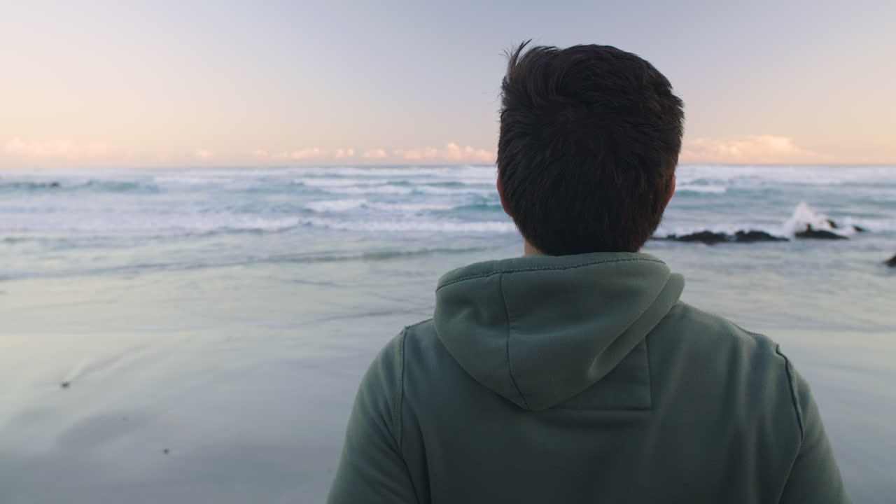 Camera orbits around young man in jogging hoody as he turns and looks at ocean before early morning run, as the sun rises over the horizon