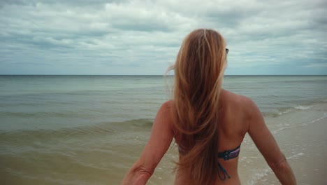 laughing, playful mature woman in bikini and sunglasses turns around and runs on the beach