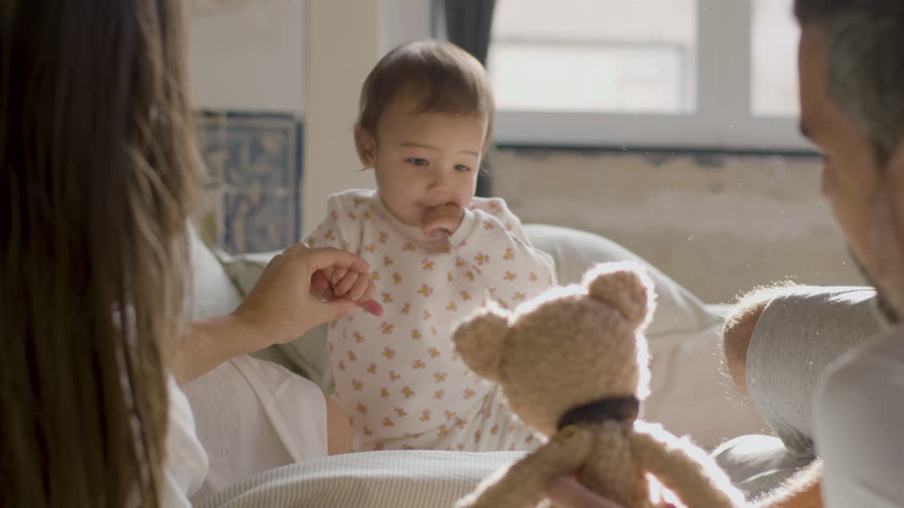 Happy Parents In Bed On Sunday Morning Playing With Their Cute Baby Girl Using A Teddy Bear