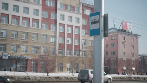 city street with tall buildings, cars moving both directions past bus stop sign on pole, winter snow along curb, clear sky and power lines overhead, urban commute scene with light traffic