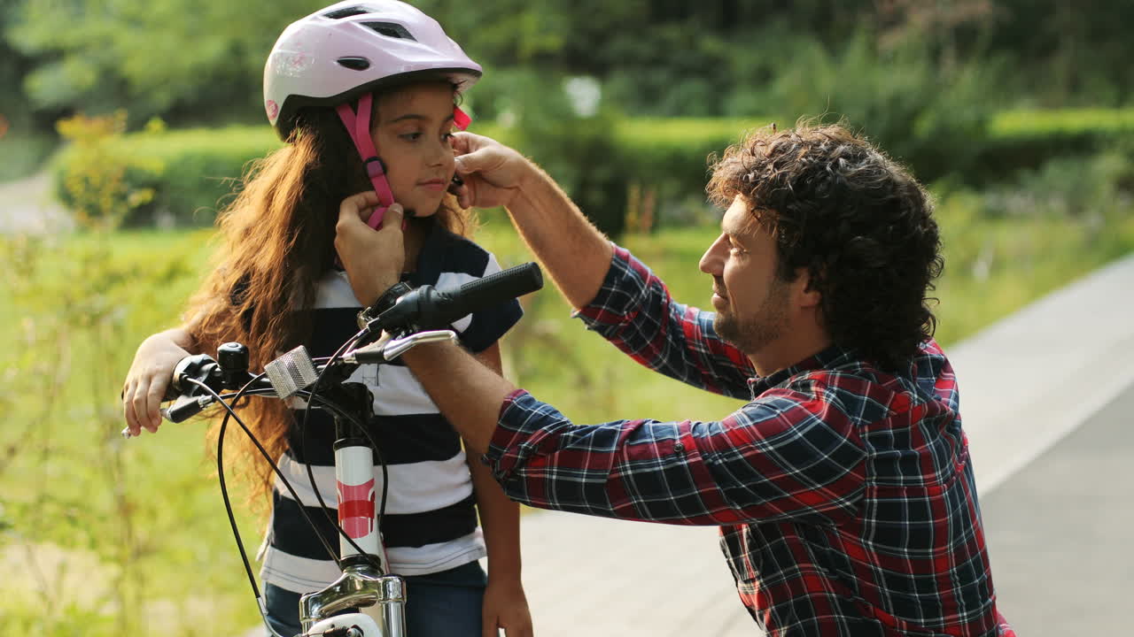 Portrait of a little girl and her father near the bike. Dad wears a helmet on the girl's head. They look at each other, then - into the camera. Smiling. Blurred background