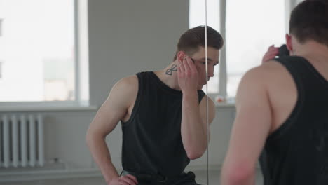 cute boy in black vest applies makeup with sponge before large mirror in bright studio, urban building visible through window, soft natural light and subtle reflection guide careful grooming