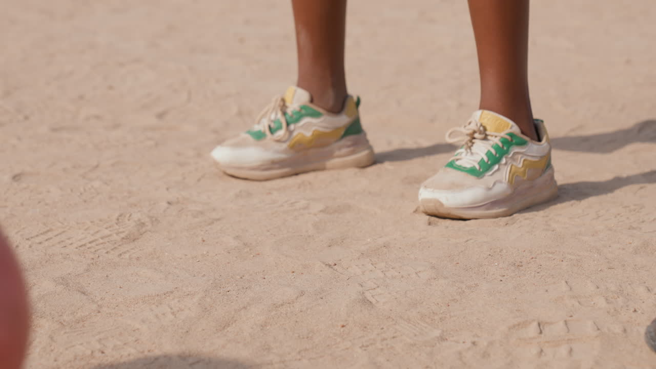 Primo piano di scarpe da ginnastica su un campo di terra, un aspirante attaccante pronto a calciare, scarpe da ginnastica verdi e bianche usurate, un pallone rosa che entra nell'inquadratura, luce solare calda e texture del terreno polveroso, focus al rallentatore sul gioco di gambe.