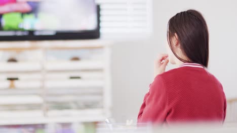 Caucasian-woman-sitting-on-couch,-eating-snacks-and-watching-tv-in-living-room