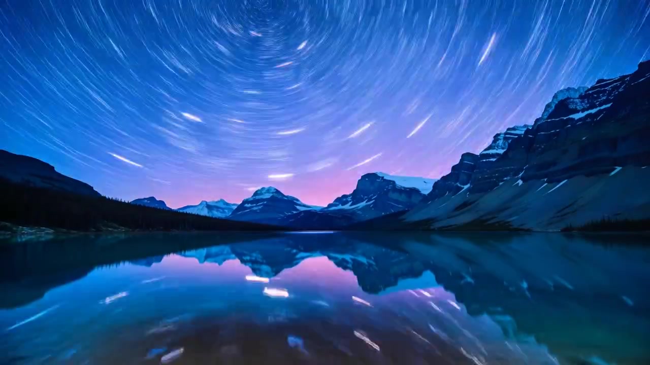 日の出が近づき、星の軌跡が残る夜明けの空の下、高山とそれを映す湖が写し出されている。