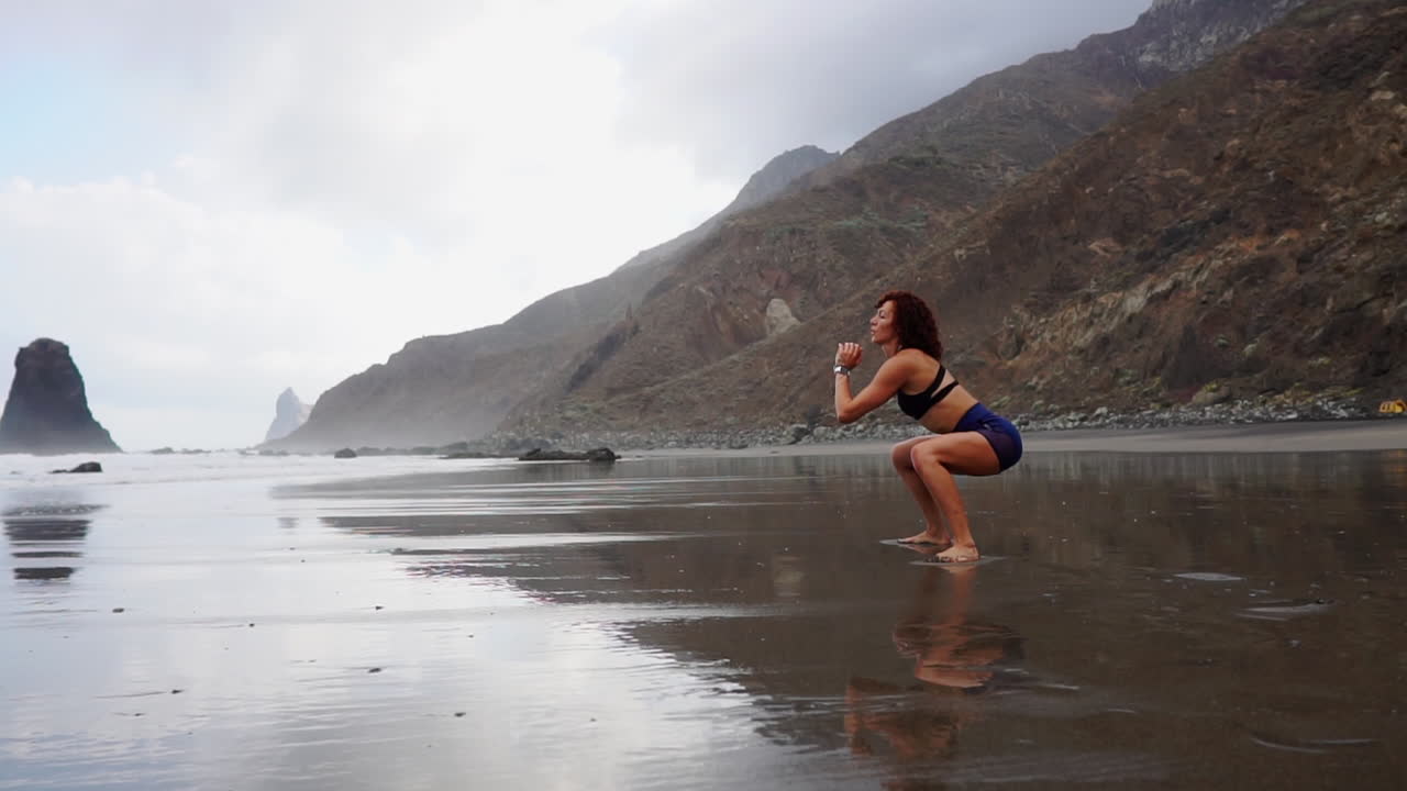 Performing squats and jumps by the ocean's shore, a young, slim woman's training is depicted in slow motion