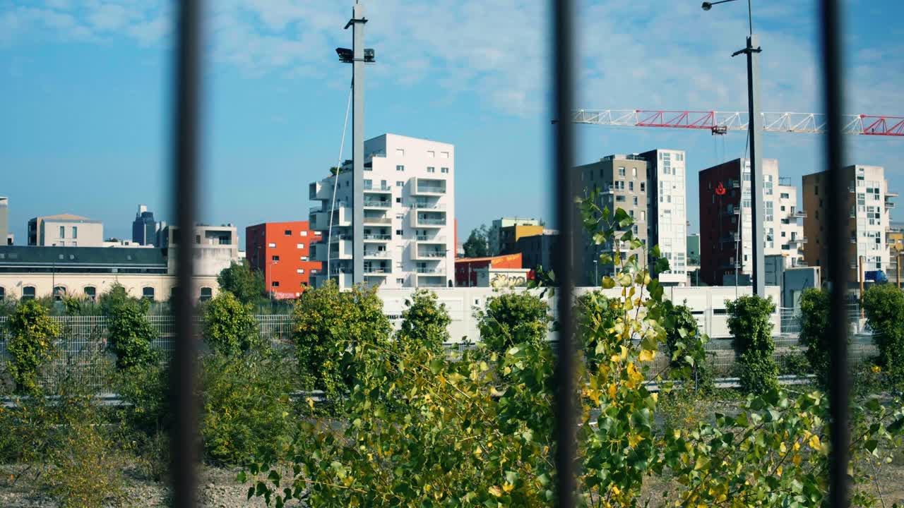 Vista attraverso una recinzione metallica che mostra nuovi edifici residenziali colorati e gru edili. Pianificazione urbana ed espansione di un quartiere cittadino con architettura e vegetazione miste