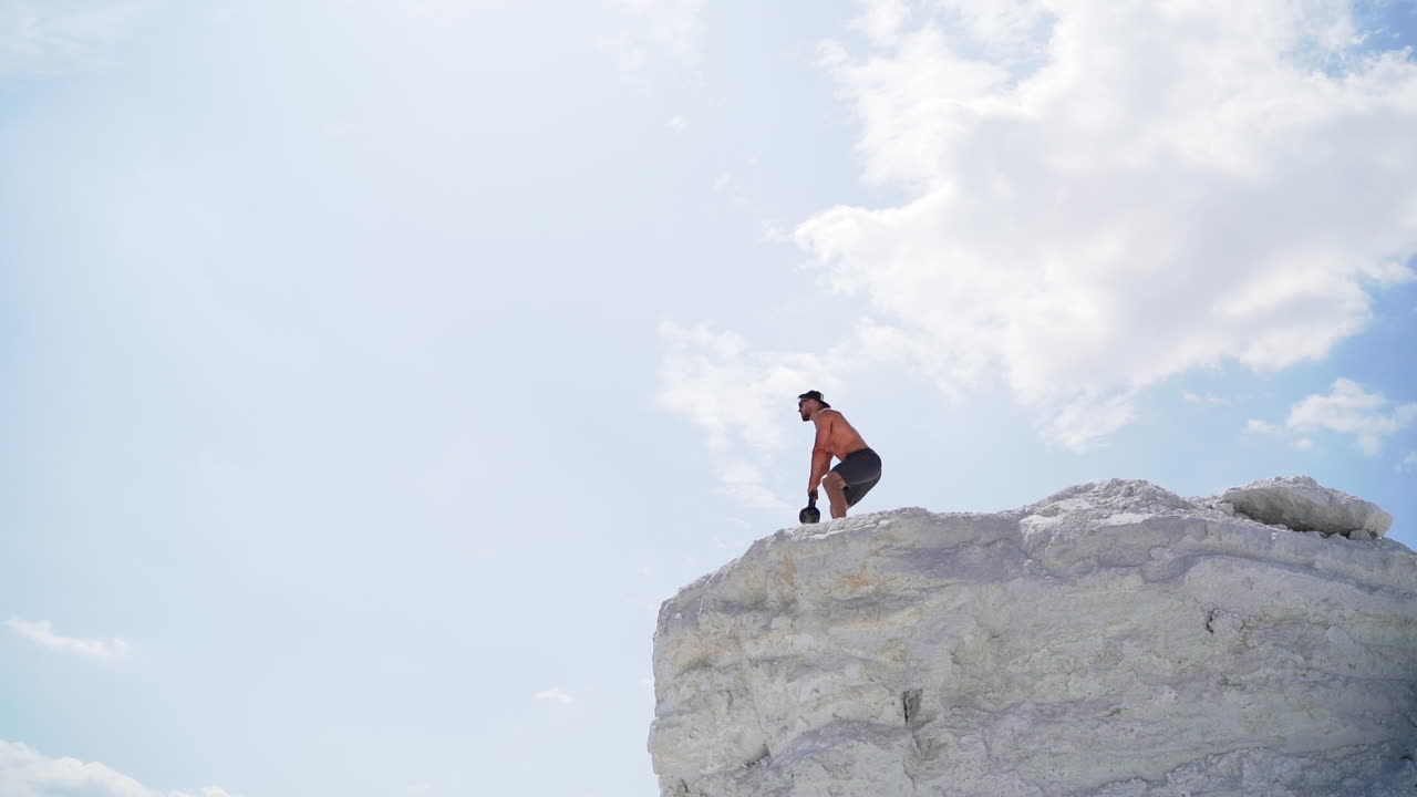 View from below on a healthy man training with heavy weight on a hill. Bodybuilder lifting kettlebell on the natural sky background. Shirtless man during workout outdoors.