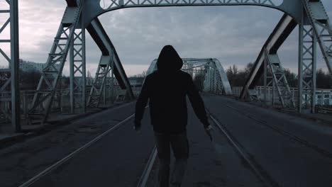 young man in hoodie and balaclava with red burning signal flare on the road under an old steel frame bridge, slow motion