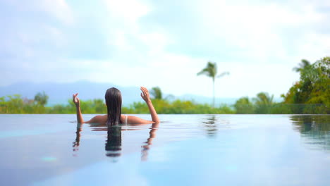 a woman in a swimming pool with her back to the camera smooths her wet hair as she looks out over a tropical paradise