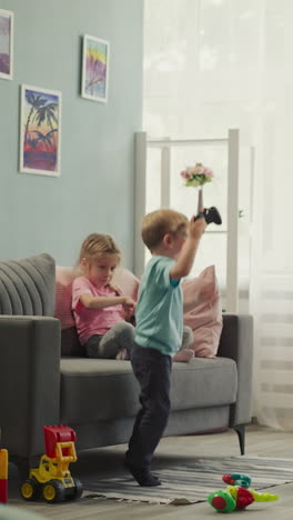 active little boy jumps while playing video game with elder sister at home on weekend. kids have fun in living room with scattered toys on floor