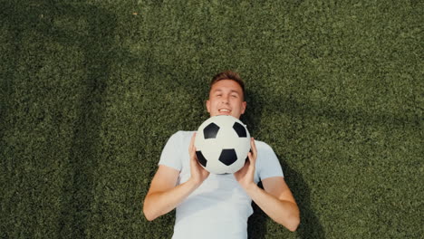 vista superior de un joven futbolista feliz tirado en el campo, lanzando y atrapando la pelota durante la sesión de entrenamiento