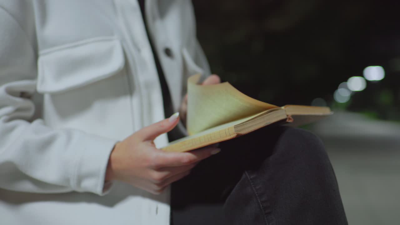 Partial view of woman seated outdoors at night gently turning page of book resting on knee while soft bokeh lights glow in background and surrounding trees