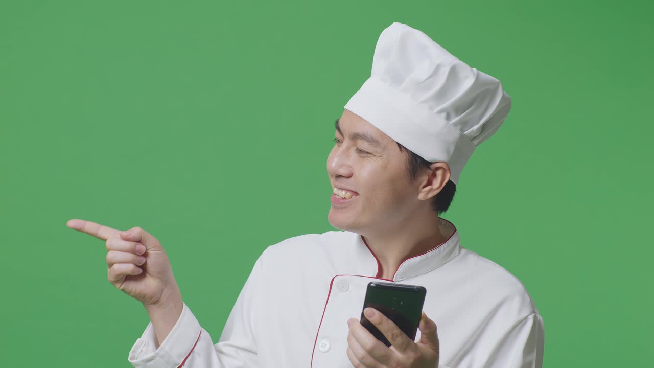 Close Up Of Asian Man Chef Using Smartphone And Pointing To Side While Standing In The Green Screen Background Studio
