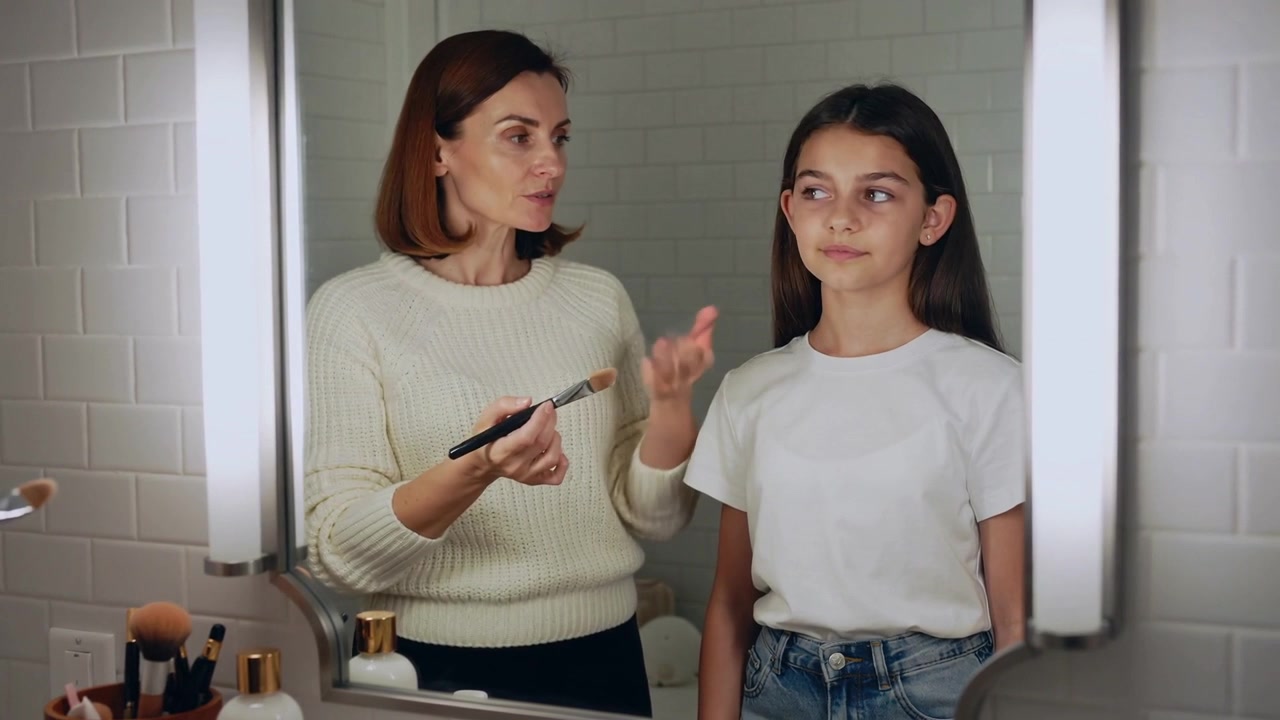 Donna che si protende, con un pennello, trucca una ragazza davanti allo specchio del bagno con illuminazione a LED, insegnando.